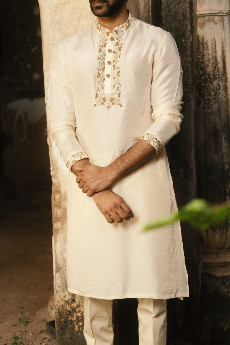 Man wearing a white kurta with intricate embroidery standing against a textured wall.