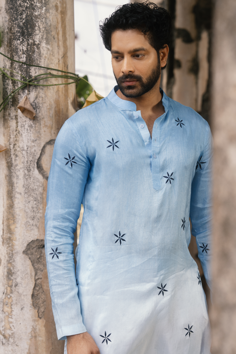 Man wearing a light blue kurta with star patterns against a rustic wall.