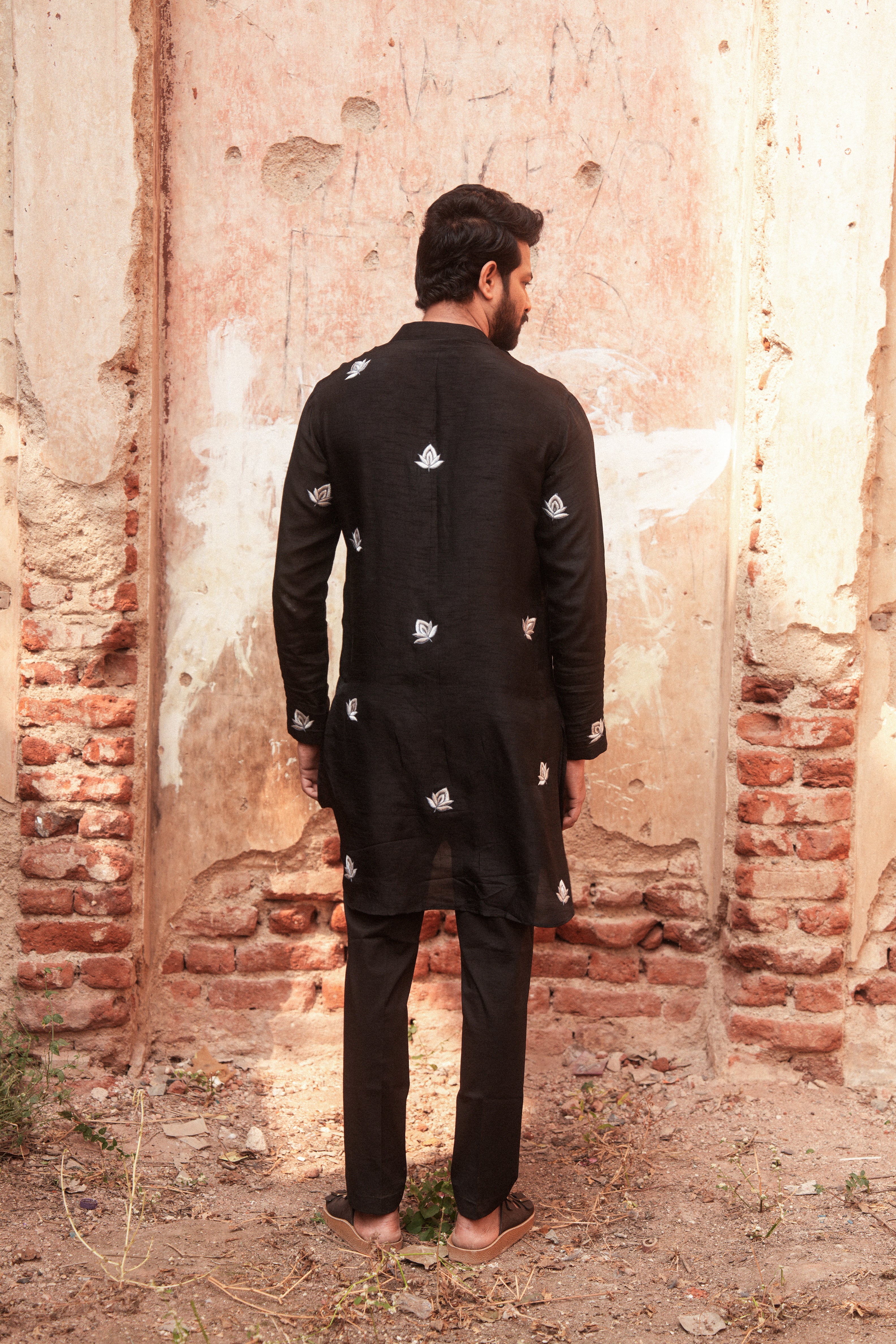 Man wearing a black traditional outfit with white patterns standing against a rustic brick wall.