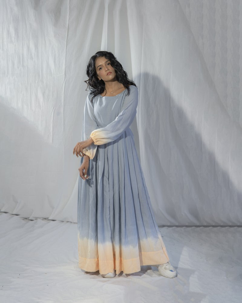 Woman wearing a long, light blue and beige dress against a white curtain background